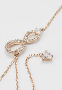 Colar em tom de ouro em forma de infinito, apresentando strass claros, um design texturizado e uma corrente delicada com um destaque de pedra triangular.