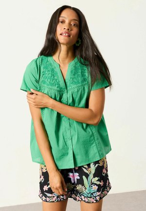 Frau mit langen dunklen Haaren, die eine grüne bestickte Bluse mit kurzen Ärmeln und schwarze Shorts mit Blumenmuster trägt, steht mit verschränktem Arm.