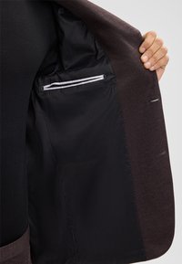 Blazer marrone con una superficie liscia e interno nero. Presenta una tasca con una rifinitura bianca sul lato sinistro, con cuciture interne visibili.