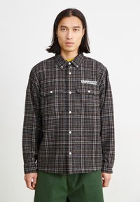 Chemise à carreaux noir et marron avec manches longues, col button-down, deux poches poitrine et détail de logo blanc sur le côté gauche.
