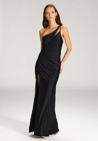 Robe noire à une épaule en tissu lisse et extensible. Présente un détail noué à la taille et une fente haute sur la cuisse pour une élégance supplémentaire.