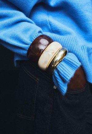 Ręka w niebieskim, dzianinowym swetrze i ciemnych dżinsach z trzema masywnymi bransoletkami w odcieniach brązu, kremu i złota na nadgarstku.