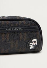 Czarna kosmetyczka z teksturowanym wykończeniem, brązowym wzorem logo oraz gumowym akcentem przedstawiającym postać w okularach przeciwsłonecznych z kotem.