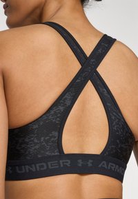 Black athletic bra featuring crisscross straps, textured fabric, and a supportive underband with the logo "UNDER ARMOUR" in dark lettering.