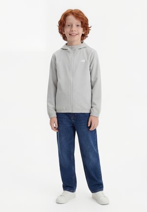 Rødhåret barn smilende, iført lysegrå lynlås-hættetrøje, blå jeans og hvide sneakers, stående mod en ensfarvet hvid baggrund.