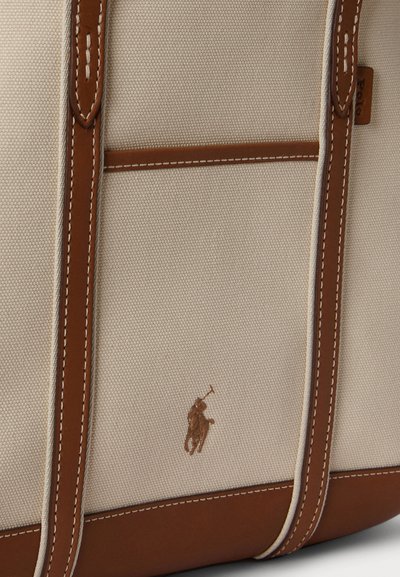Τσάντα ώμου από καμβά με καφέ δερμάτινες λεπτομέρειες, με ενισχυμένες ραφές και μικρό, ανάγλυφο λογότυπο. Κρεμ φόντο με αντιθετικές πινελιές.