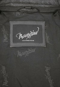 Schwarzes Stoffetikett mit "Mauritius Leatherwear" in weißer Schrift. Weiche Textur, mit sich wiederholendem "Mauritius"-Muster in helleren Schwarz.