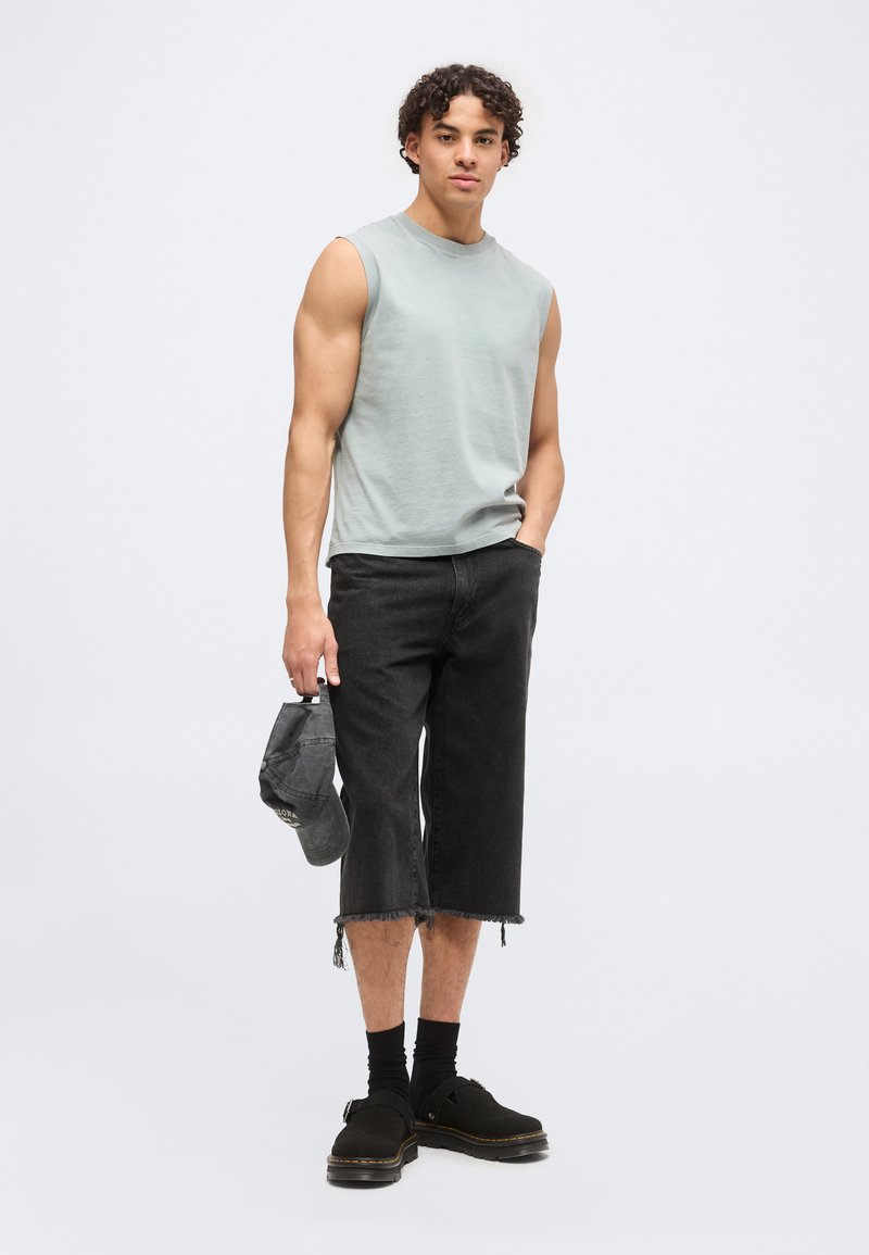 Joven con cabello rizado que lleva una camiseta sin mangas de color gris claro, vaqueros negros cortos, calcetines negros, zapatos negros, sosteniendo una gorra negra en una mano.