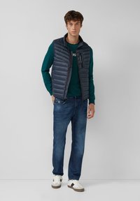 Gepolsterte marineblaue Weste mit hohem Kragen, mit Reißverschluss versehene Fronttasche, getragen über einem grünen Langarmshirt und kombiniert mit blauen Jeans und weißen Turnschuhen.