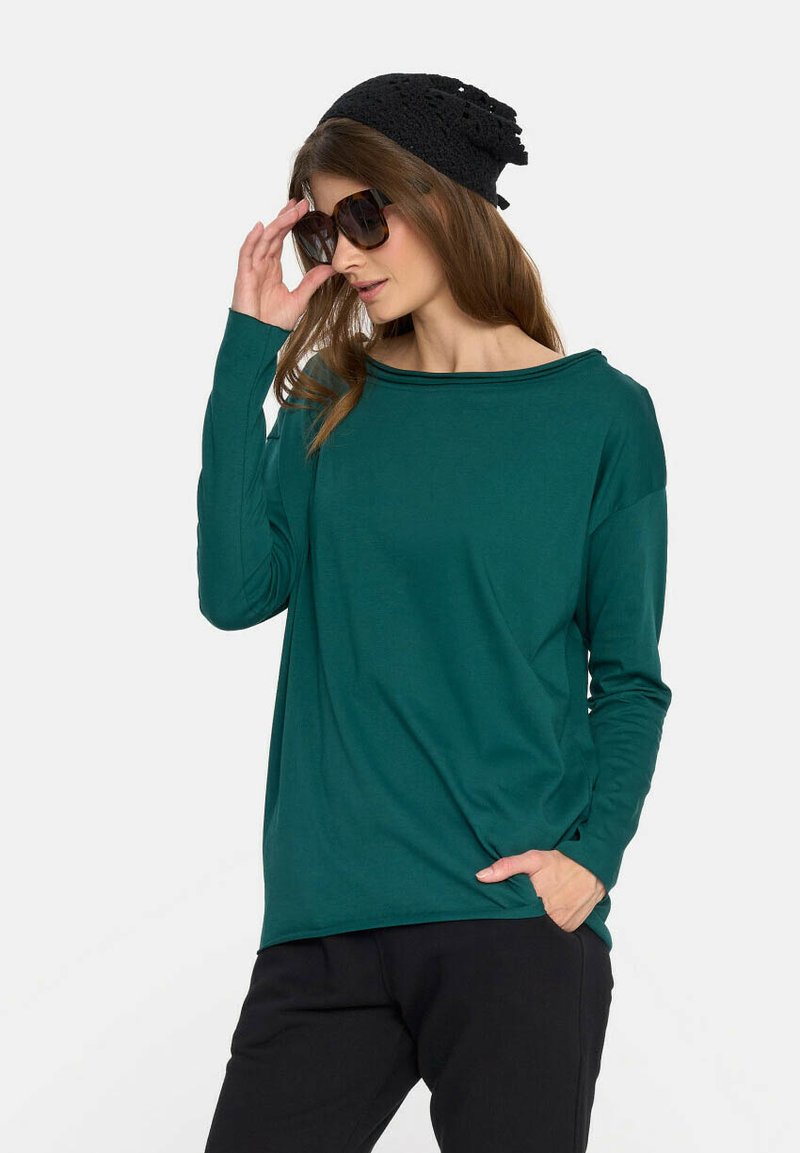 Turkusowa koszula z długim rękawem i szerokim dekoltem, w luźnym kroju, zestawiona z czarnymi spodniami. Modelka nosi czarną czapkę i oversize'owe okulary słoneczne.