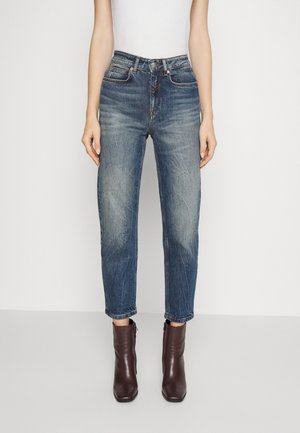 Vrouw die versleten blauwe high-waist jeans en donkerbruine enkellaarzen draagt, staand met ontspannen handen langs het lichaam op een lichte achtergrond.