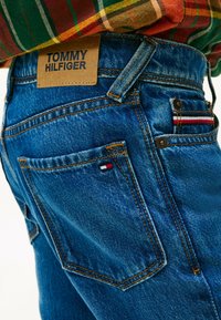 Tmavě modré džíny z denimu s koženou nášivkou TOMMY HILFIGER. Obsahují zadní kapsy se červenobílými a modrými akcenty a kontrastním oranžovým prošíváním.