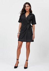 Black wrap dress with short sleeves, a tied waist detail, contrasting white stitching, and a collar. Worn with black high-heeled shoes.
