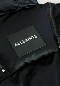 Svart nylonjacka med blank yta, sidodragkedja och fyrkantig svart etikett med präglad vit text "ALLSAINTS."