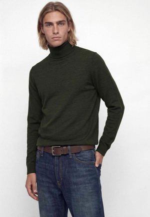 Homme portant un pull à col roulé vert foncé, un jean bleu et une ceinture marron, debout devant un fond blanc uni.