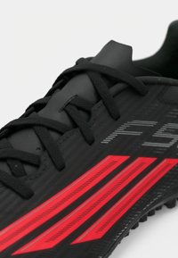 Κοντινό πλάνο μαύρου αθλητικού παπουτσιού με μαύρες κορδόνες και τρεις έντονες κόκκινες ρίγες στο πλάι σε απλή φόντο.