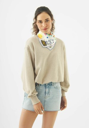Femme portant un pull beige, une jupe en jean clair et un foulard blanc au cou avec un imprimé Snoopy et soleil, debout sur un fond blanc.
