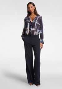 Pantaloni lunghi blu con un taglio sartoriale, abbinati a una blusa di seta multicolore con colletto arricciato e motivi astratti.