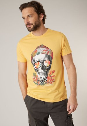 Man met baard die een geel t-shirt draagt met een kleurrijke schedelafbeelding en de tekst '"Chill"', gecombineerd met donkere cargobroek, staand met één hand in de zak.