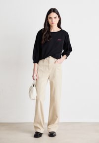 Maglietta nera a maniche lunghe con testo ricamato rosa, abbinata a pantaloni beige a gamba larga, che tiene una borsa bianca, indossando scarpe nere.