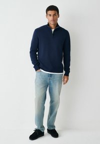 Navy Zip-up-Pullover aus Strickstoff, kombiniert mit hellblauen Jeans und schwarzen Wildlederschuhen. Schlichtes Design mit gerippten Bündchen.