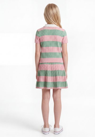 Παιδί με μακριά ξανθά μαλλιά, φοράει ένα ριγέ ροζ και πράσινο πλεκτό φόρεμα με κοντά μανίκια και λευκά αθλητικά παπούτσια, στέκεται με την πλάτη στραμμένη.