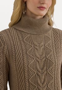 Kvinna iklädd en beige kabelstickad polotröja och hängande guldörhängen med kristalldetaljer.