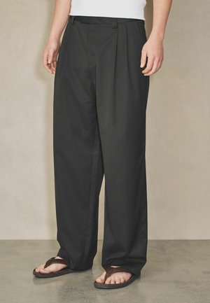 Persoană care poartă pantaloni largi negri plisati și sandale maro, stând pe un podea netedă, în fața unui perete bej.