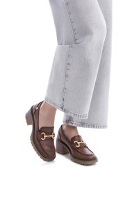 Mocasines de cuero marrón con detalle de cadena dorada, con un tacón grueso y suela de goma texturizada, combinados con jeans de pierna ancha de color gris claro.