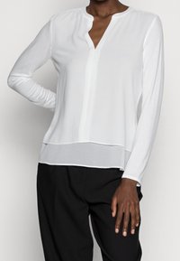 Blouse blanche à manches longues avec un décolleté en V, présentant un panneau horizontal contrastant à l'ourlet et une texture lisse. Portée avec un pantalon noir.