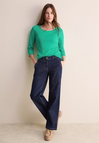 Grön långärmad topp med rundad halsringning, kombinerad med mörkblå vida jeans. Jeansen har framfickor och synlig sömnad.