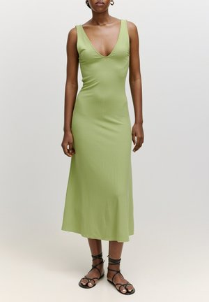 Groene geribbelde midi-jurk met een diepe v-hals en een aansluitende silhouette, aangevuld met zwarte sandalen met bandjes. Soepele structuur en minimalistisch ontwerp.