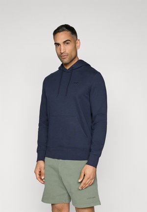 Felpa blu navy realizzata in morbido tessuto, con tasca frontale, cappuccio regolabile con coulisse e un piccolo logo sul petto. Il modello indossa pantaloni corti beige.