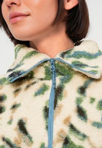 Chaqueta de forro polar con cierre de cremallera, textura suave, fondo crema con patrones abstractos en verde y marrón, y cremallera azul.