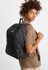 Μαύρος σακίδιο πλάτης με επενδυμένο σχέδιο, κίτρινη ραφή και ρυθμιζόμενους ιμάντες. Διαθέτει μπροστινή τσέπη και ελαστικό κορδόνι για επιπλέον αποθήκευση.