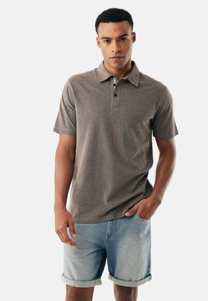 Mann trägt ein braunes, kurzärmeliges Poloshirt und hellblaue Jeansshorts, eine Hand in der Tasche, vor einem einfarbigen Hintergrund.