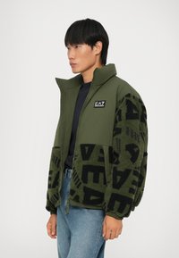 Giacca verde con design in patchwork che presenta un tessuto nero peloso e motivi di logo audaci. Collo alto e chiusura con zip; vestibilità casual.
