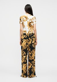 Γυναίκα με μακριά σκούρα μαλλιά φοράει λευκό κοντομάνικο μπλουζάκι με χρυσό μπαρόκ σχέδιο και μαύρο παντελόνι φαρδιάς γραμμής με αντίστοιχο χρυσό μπαρόκ μοτίβο.