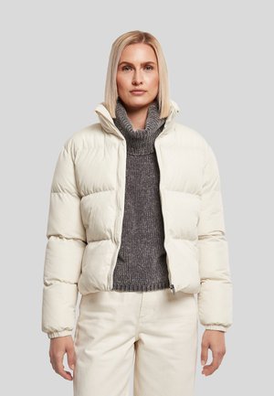 Femme portant une doudoune crème sur un pull à col roulé gris avec un pantalon crème, debout devant un fond clair uni.