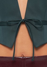 Nahaufnahme des mittleren Körperbereichs mit einem dunkelgrünen, bauchfreien Oberteil, das über freiliegender Haut oberhalb von burgunderroten Lederhosen zu einer Schleife gebunden ist.