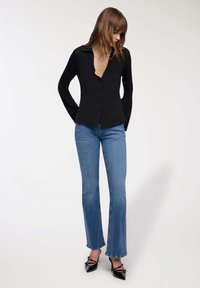 Donna con capelli castani che indossa una camicia nera con bottoni, jeans blu a gamba dritta e tacchi alti neri a punta, in piedi davanti a uno sfondo neutro.