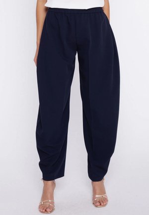 Kobieta w luźnych granatowych spodniach z elastycznym pasem i złotych sandałach na cienkich paskach, na białym tle.