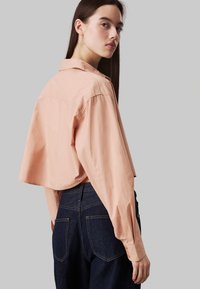 Camicetta corta rosa chiaro con colletto, polsini abbottonati e una texture liscia, abbinata a jeans in denim scuro con tasche a toppe.