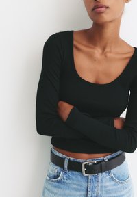 Schwarzes geripptes Langarmoberteil mit U-Ausschnitt, kombiniert mit hoch taillierten hellblauen Jeans und einem schwarzen Gürtel mit einer silbernen Schnalle.