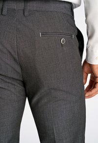 Pantaloni grigi da abito con una texture fine, caratterizzati da una tasca posteriore con dettaglio a bottone e un design sagomato. La cintura è dritta e ordinata.