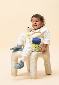 Baumwolloverall in Creme mit bunten Applikationen (gelb, blau, grün). Sitzt auf einem gepunkteten weißen Hocker, trägt blaue Schuhe und graue Socken.