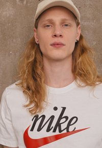 Maglietta bianca con logo "nike" in nero e rosso. Capelli lunghi e ondulati, indossa un cappellino beige. Sfondo semplice con un muro di cemento testurizzato.