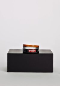 Pomada do włosów w okrągłej puszce z pomarańczową pokrywką, czarną podstawą i czerwoną etykietą z napisem "Uppercut Deluxe". Gładka tekstura, kompaktowy kształt.