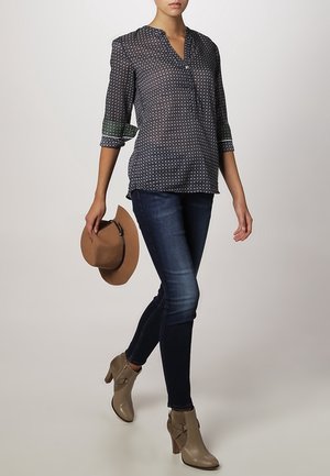 Femme portant un chemisier à motifs, un jean skinny foncé, des bottines taupe, tenant un chapeau à large bord marron dans sa main droite.