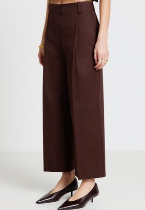 Femme portant un pantalon marron foncé taille haute et à jambes larges, associé à des chaussures noires à bout pointu et talon bas, ainsi qu'à des bijoux dorés aux mains.